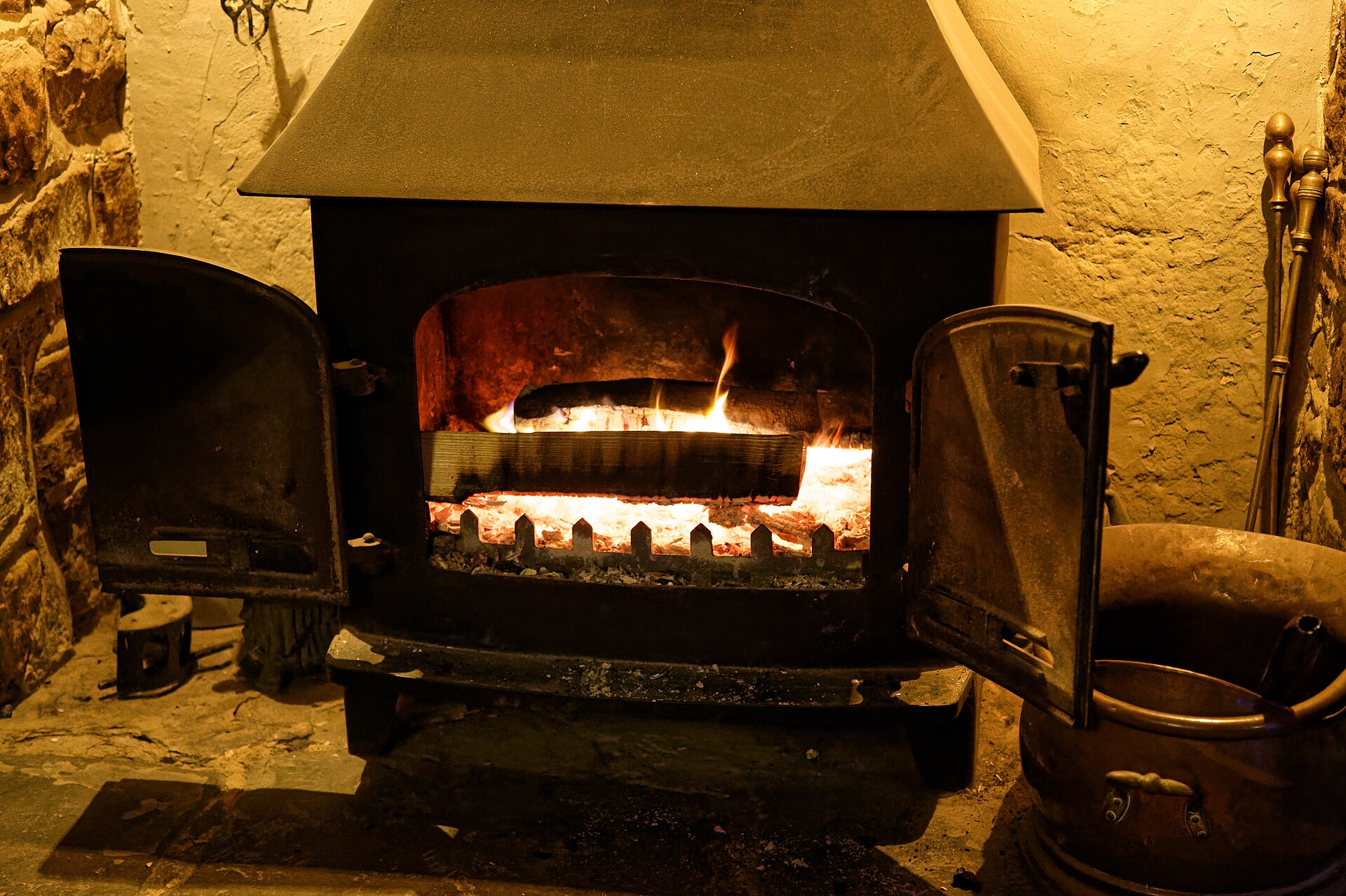 A black wood-burning stove with both doors open and a log burning inside, beside a copper coal scuttle and fire tools.